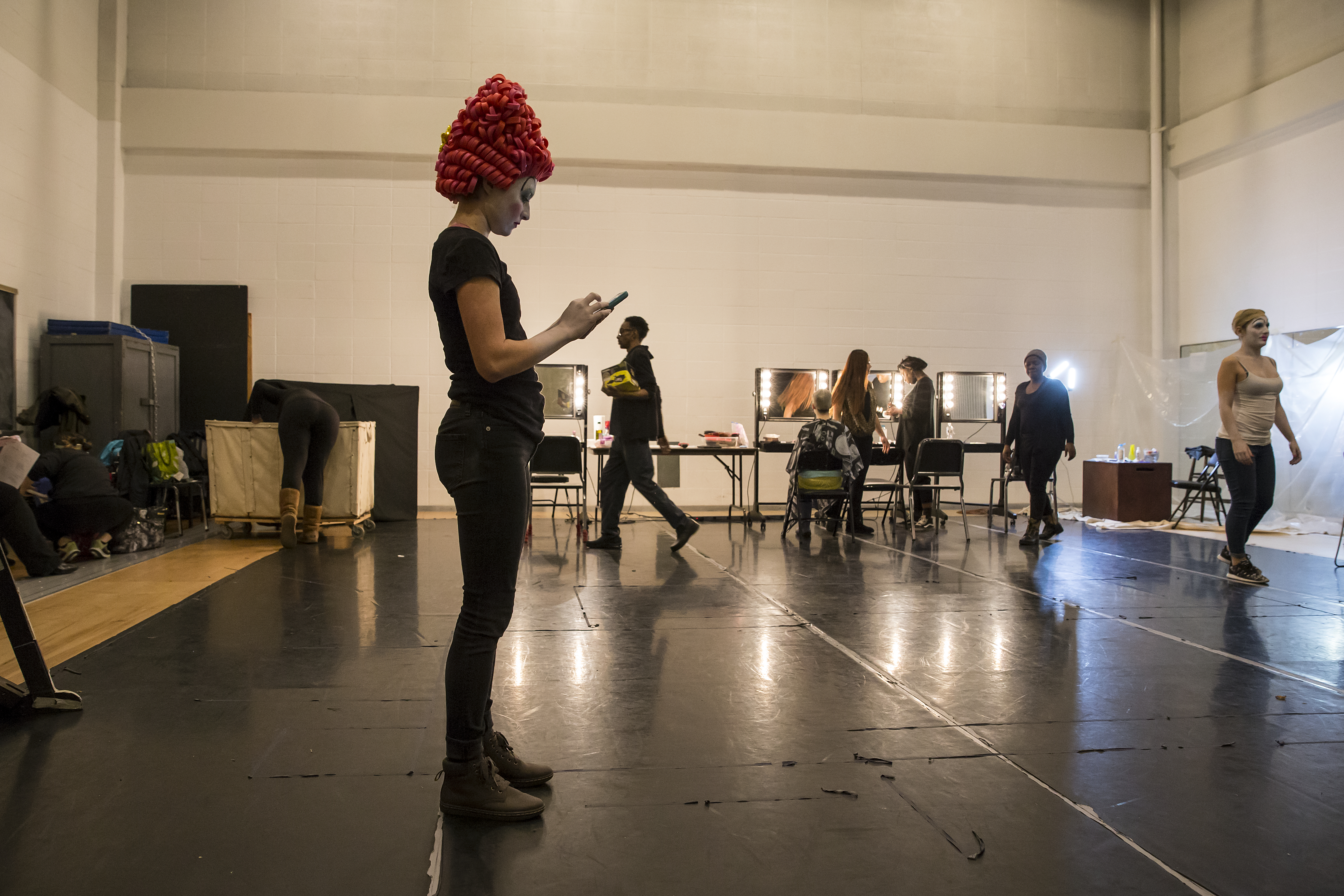 Large backstage room with student in hair and makeup looking at phone, other students in background