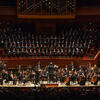 Orchestra and large choir performing on stage with pipe organ above choir. 