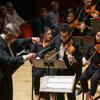 Violinists performing in an orchestra with male conductor and audience behind them.