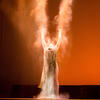 Performer with arms raised above head against an orange backdrop.